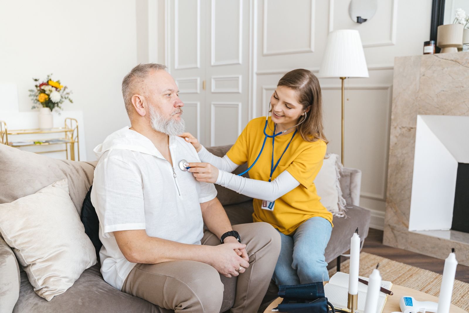 A home health nurse checking a patient's vitals with a stethoscope in a comfortable living room